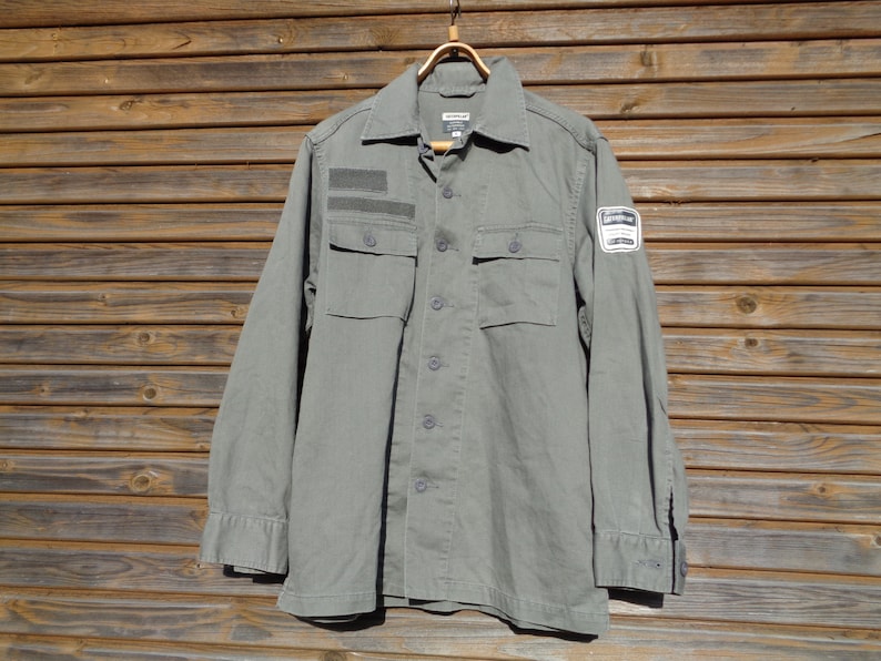 Puede incluir: Una camisa verde de manga larga con dos bolsillos y un parche en la manga derecha. El parche tiene el texto "WORKWEAR".