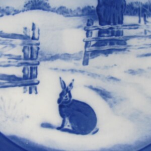 May include: Blue and white ceramic plate with a winter scene featuring a rabbit sitting in the snow near a wooden fence.