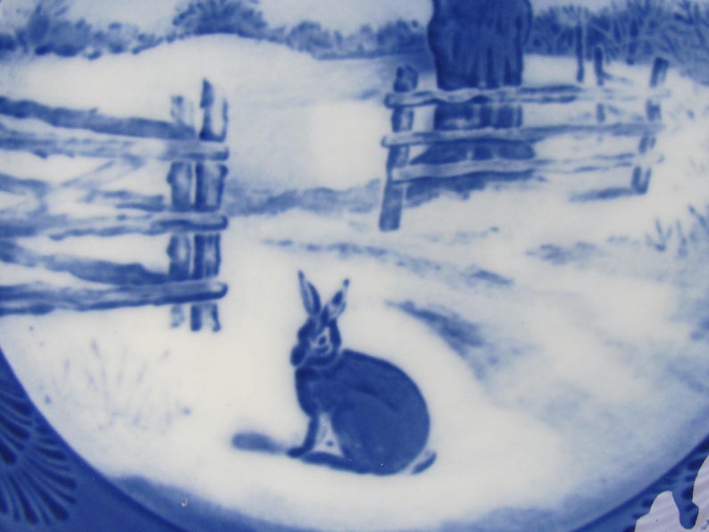 May include: Blue and white ceramic plate with a winter scene featuring a rabbit sitting in the snow near a wooden fence.