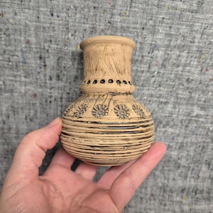 May include: A small, beige ceramic vase with a bulbous body and a flared rim. The vase features a series of horizontal lines around the base and floral patterns. The vase is held in a hand.