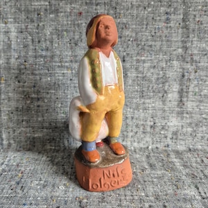 May include: A ceramic figurine of a person in a traditional outfit. The figure wears a brown hat, a green vest over a white shirt, and yellow pants. The figurine is standing on a brown base with the text "Nils Holgersson".