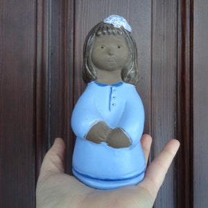 May include: A ceramic figurine of a girl with brown hair and a blue dress. She is wearing a flower crown and has a neutral expression on her face.