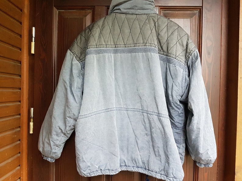 May include: A blue and grey quilted jacket with a zip closure. The jacket has a diamond pattern on the upper back and a solid colour on the lower back.
