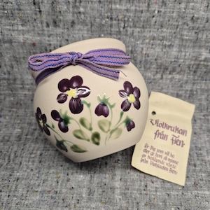 May include: A small, cream-colored ceramic pot with hand-painted purple flowers and green leaves. A purple ribbon is tied around the top. A small tag with text is attached to the pot.