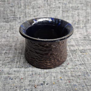 May include: A small, round ceramic bowl with a dark brown, textured exterior and a smooth, glossy deep blue interior. The rim is wide and slightly flared, featuring a dark, reflective finish that contrasts with the rough, earthy texture of the body.