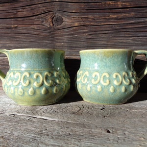 May include: Two green ceramic mugs with a light green glaze and a raised design of circles and teardrops. The mugs are sitting on a wooden surface.
