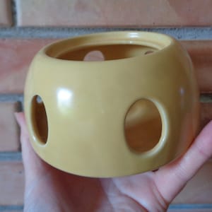 Könnte beinhalten: Ein gelber Keramiktopf mit drei runden Löchern. Der Topf ist glatt und hat eine glänzende Oberfläche.