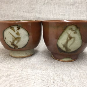 May include: Two brown ceramic teacups with white glaze and brown calligraphy designs. The cups are sitting on a beige fabric surface.