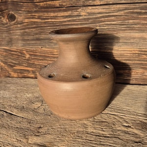 Könnte beinhalten: Eine handgefertigte, braune Keramikvase mit bauchigem Boden und schmalem Hals. Die Vase hat mehrere kleine Löcher in der Nähe der Oberseite, wahrscheinlich zum Anordnen von Blumen. Das rustikale Design und die Erdtöne deuten auf eine natürliche, organische Ästhetik hin.
