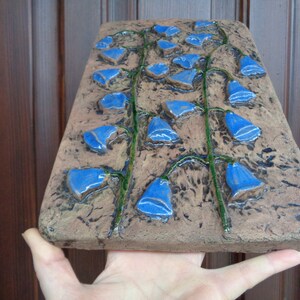 K&ouml;nnte beinhalten: Ein Wandbehang aus Keramik mit braunem Hintergrund und blauen Blumen. Die Blumen sind glockenf&ouml;rmig und vertikal angeordnet.