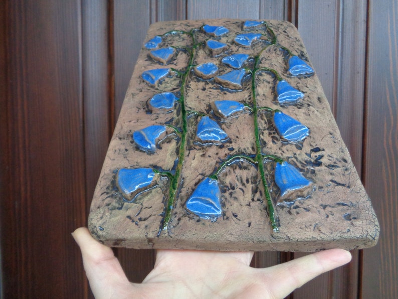 K&ouml;nnte beinhalten: Ein Wandbehang aus Keramik mit braunem Hintergrund und blauen Blumen. Die Blumen sind glockenf&ouml;rmig und vertikal angeordnet.
