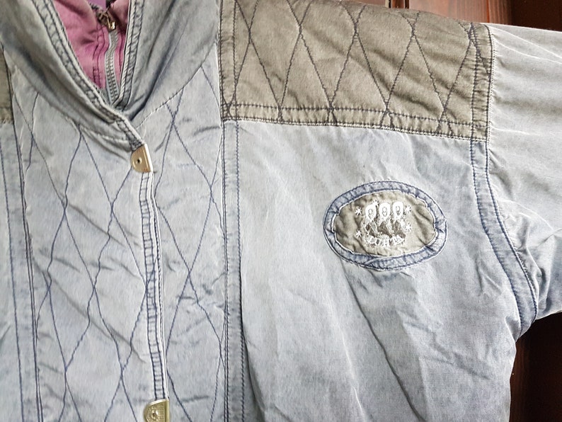 May include: A blue quilted jacket with a brown quilted patch on the shoulder. The jacket has a zip fastening and a button on the front. The jacket has a logo patch on the right sleeve that says "Pepe Jeans London".