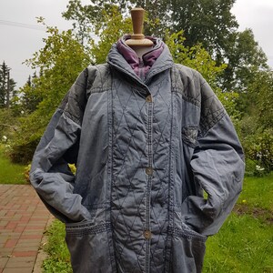 May include: A grey quilted jacket with a purple lining and a zip closure. The jacket has a drawstring at the waist and two pockets on the front.