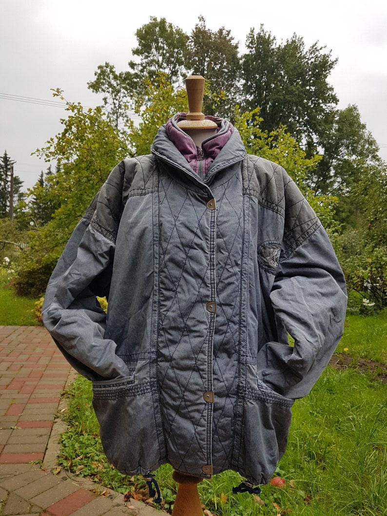 May include: A grey quilted jacket with a purple lining and a zip closure. The jacket has a drawstring at the waist and two pockets on the front.