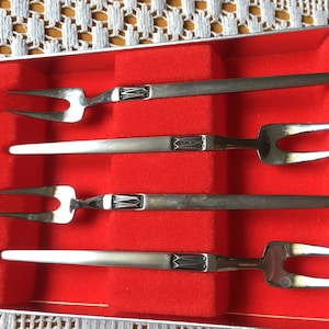 Könnte beinhalten: Satz mit vier silberfarbenen Cocktailgabeln in einer mit rotem Samt ausgekleideten Schachtel. Die Gabeln haben ein dekoratives Design am Griff.