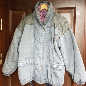 May include: A grey quilted jacket with a purple lining and a black patch on the left sleeve. The jacket has a zip closure and a drawstring at the bottom.