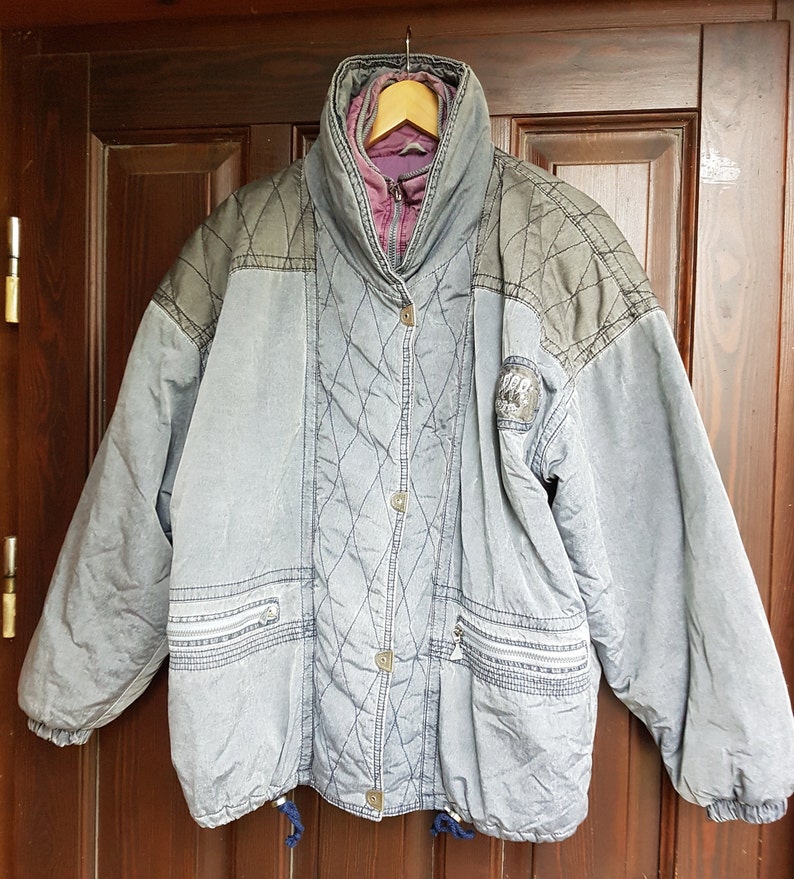 May include: A grey quilted jacket with a purple lining and a black patch on the left sleeve. The jacket has a zip closure and a drawstring at the bottom.