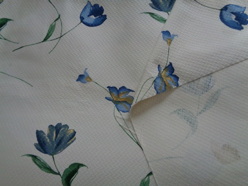 May include: White fabric with a blue and yellow floral pattern. The fabric has a textured surface and the flowers are stylized.