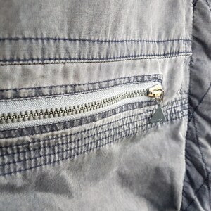 May include: Close-up of a grey zipper pocket on a garment. The zipper is silver and the pocket is outlined with dark blue stitching.
