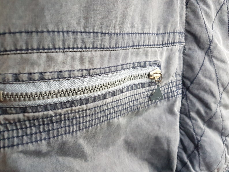 May include: Close-up of a grey zipper pocket on a garment. The zipper is silver and the pocket is outlined with dark blue stitching.