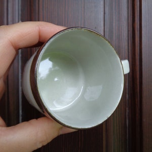 Puede incluir: Una taza de caf&eacute; de cer&aacute;mica blanca con una banda y borde marr&oacute;n. La taza tiene un asa peque&ntilde;a y un interior verde claro. La taza est&aacute; sostenida en la mano de una persona.