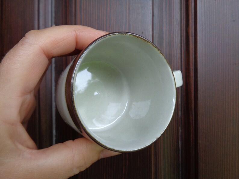 Puede incluir: Una taza de caf&eacute; de cer&aacute;mica blanca con una banda y borde marr&oacute;n. La taza tiene un asa peque&ntilde;a y un interior verde claro. La taza est&aacute; sostenida en la mano de una persona.