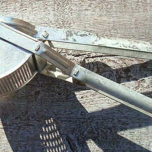 May include: A vintage, silver potato ricer with a perforated bowl and long handles. The kitchen utensil is made of metal and is designed to press cooked potatoes. The ricer is shown on a weathered wooden surface.