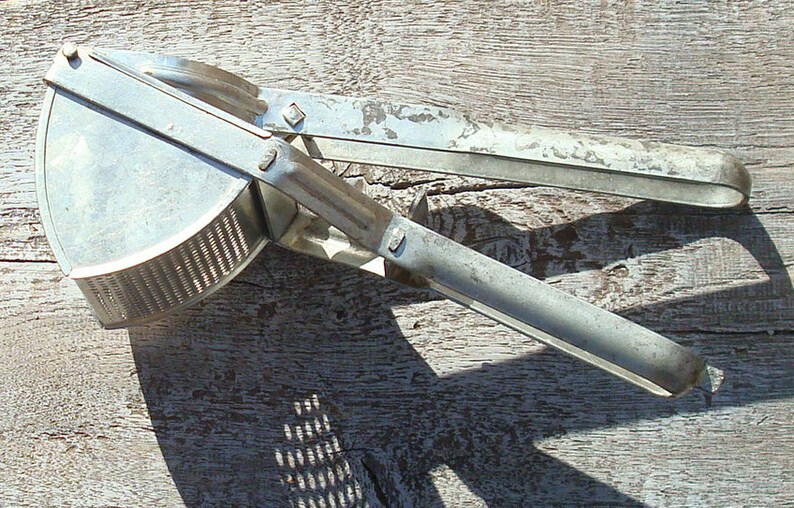 May include: A vintage, silver potato ricer with a perforated bowl and long handles. The kitchen utensil is made of metal and is designed to press cooked potatoes. The ricer is shown on a weathered wooden surface.