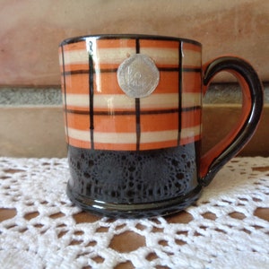 May include: A small, orange and black checkered ceramic mug with a silver coin design on the front. The mug has a black glazed bottom and a brown handle.