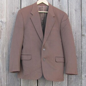 Peut inclure: Un blazer marron à deux boutons et deux poches avant est suspendu à un cintre en bois. Le blazer est fait d'un tissu de couleur unie et a un design classique et ajusté. L'arrière-plan est une surface en bois vieilli.