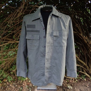 Puede incluir: Una camisa utilitaria de manga larga de color verde con un parche en el pecho izquierdo y dos bolsillos grandes en la parte delantera.