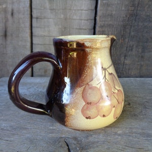 May include: A brown and cream ceramic pitcher with a handle. The pitcher has a floral design with brown leaves and orange berries.