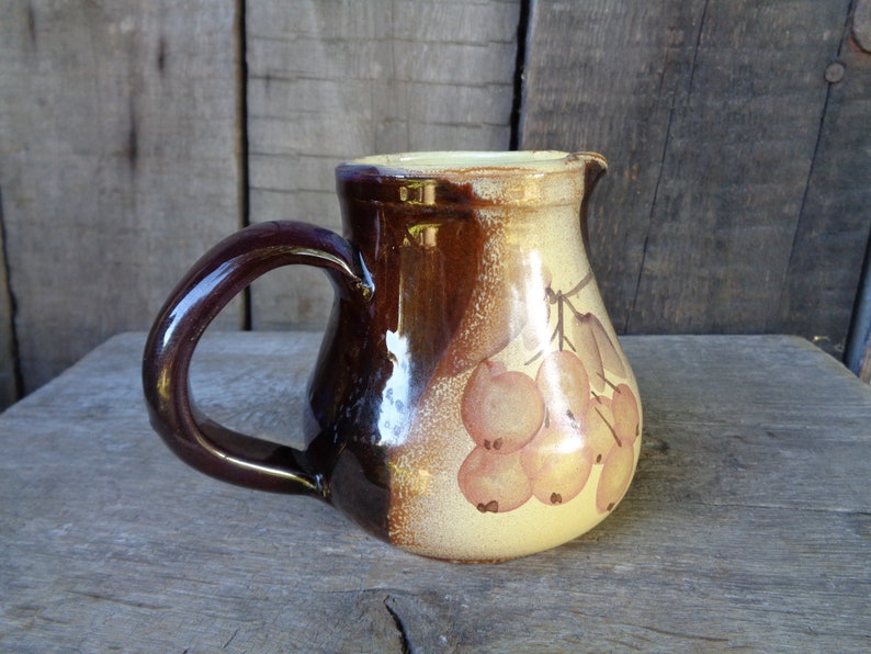 May include: A brown and cream ceramic pitcher with a handle. The pitcher has a floral design with brown leaves and orange berries.