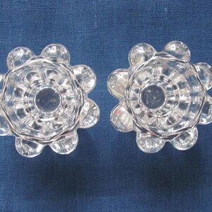 May include: Two clear glass candle holders with a flower-like design. Each holder has a central cup and eight rounded petals.