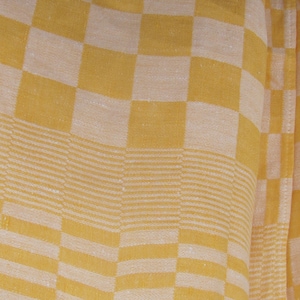 May include: A textile fabric featuring a repeating chequered pattern in shades of yellow and off-white. The material displays a woven texture with alternating blocks of colour and horizontal striped bands integrated into the design of the cloth.