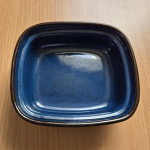 Puede incluir: Un cuenco cuadrado de cerámica con esquinas redondeadas. El interior es de un azul profundo brillante y el borde es de color marrón oscuro. El cuenco está vacío y se encuentra sobre una superficie de madera clara.