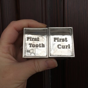 May include: Two small silver boxes with engraved text. The box on the left says "First Tooth" and the box on the right says "First Curl".