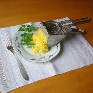 May include: A stainless steel potato ricer sits on a white plate with yellow, riced potatoes and fresh parsley. A silver fork rests on a white, textured placemat. The plate has a black and gold decorative border.