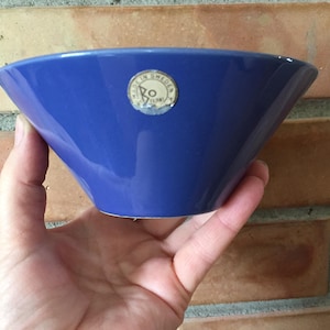 Könnte beinhalten: Eine leuchtend blaue Keramikschale mit ausgestelltem Design. Die Schale hat eine glatte, glänzende Oberfläche und einen kleinen, runden Aufkleber an der Seite. Die Schale wird in einer Hand gehalten.