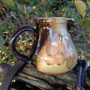May include: A brown ceramic pitcher with a handle and a painted design of a bunch of yellow berries. The pitcher is sitting on a wooden surface.