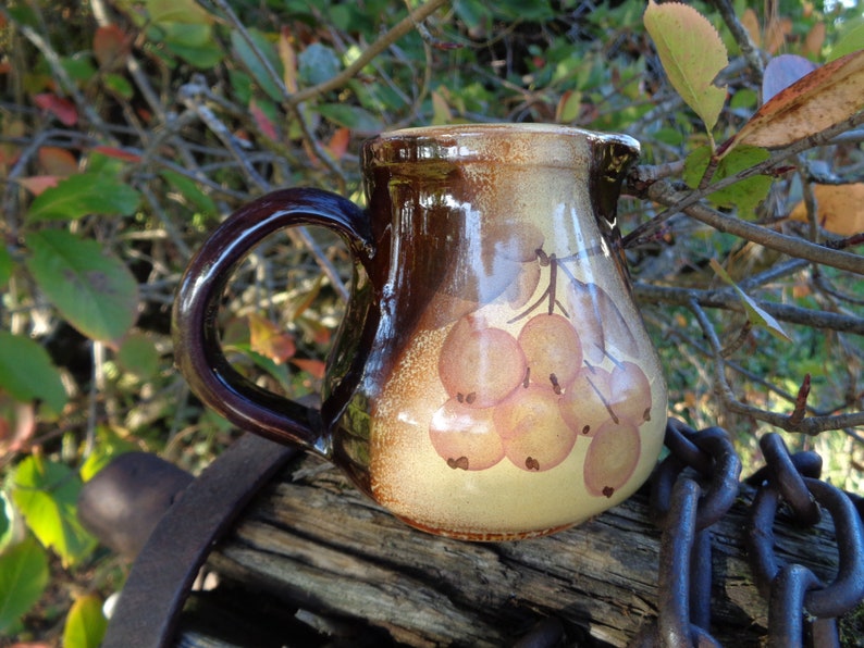 May include: A brown ceramic pitcher with a handle and a painted design of a bunch of yellow berries. The pitcher is sitting on a wooden surface.