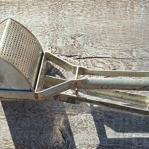May include: A vintage potato ricer in silver metal, with a perforated basket and a long handle. This kitchen utensil is designed for mashing cooked potatoes and other soft foods. The ricer is made of metal and rests on a weathered wooden surface.