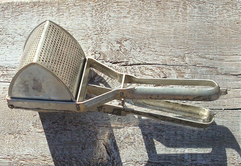 May include: A vintage potato ricer in silver metal, with a perforated basket and a long handle. This kitchen utensil is designed for mashing cooked potatoes and other soft foods. The ricer is made of metal and rests on a weathered wooden surface.