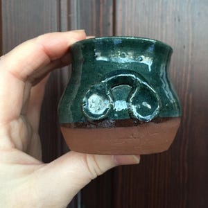 Op de afbeelding: Een kleine, groene keramische pot met een bruine rand. De pot heeft een uniek ontwerp met twee ronde bulten aan de voorkant.