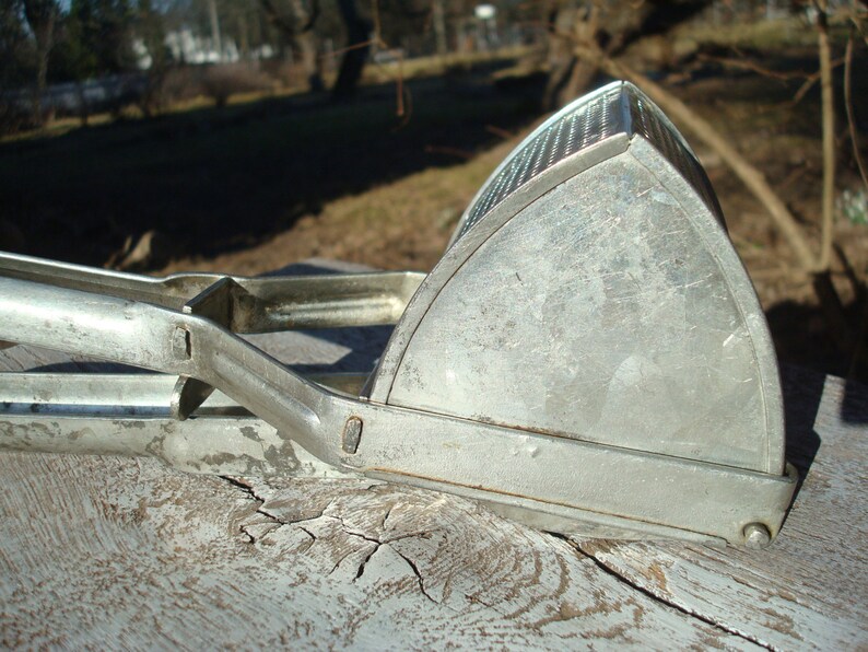 May include: A vintage, silver potato ricer. The kitchen utensil has a triangular head with a perforated surface and long handles for pressing. The ricer is made of metal and shows signs of wear, suggesting it is an antique kitchen tool.