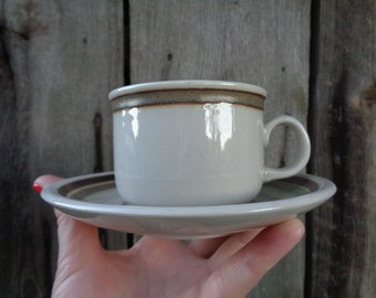 Peut inclure: Un ensemble tasse et soucoupe en céramique de couleur gris clair. La tasse présente une anse arrondie et lisse ainsi qu'une bande brun foncé sur le bord supérieur. La soucoupe assortie possède une bande circulaire brun foncé.