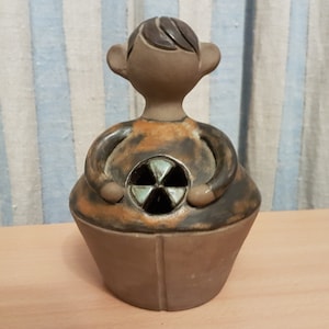 May include: A ceramic figurine of a person with a brown and orange body, brown hair, and a brown face. The figurine has a hole in the center of its body, and a green and brown design inside the hole.