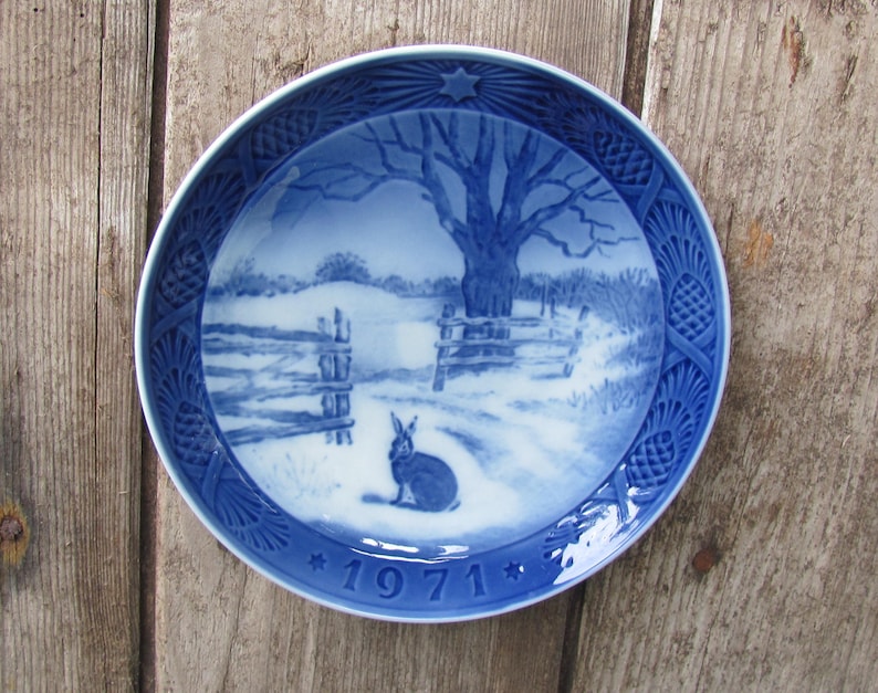 May include: A blue and white ceramic plate with a winter scene featuring a rabbit, a tree, and a fence. The plate has a blue and white border with a pine cone design. The year 1971 is printed on the bottom of the plate.