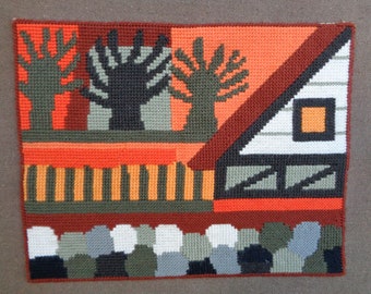 May include: A colorful needlepoint wall hanging with a geometric design featuring a house, trees, and a fence. The house is white with a red roof and a black window. The trees are green and brown. The fence is brown and orange.