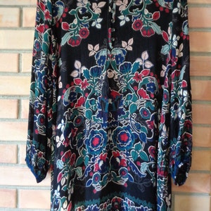 May include: Black tunic dress with a floral pattern in red, blue, teal, and white. The long-sleeved dress has a V-neckline with a tassel tie and button details. The dress is hanging on a wooden hanger.